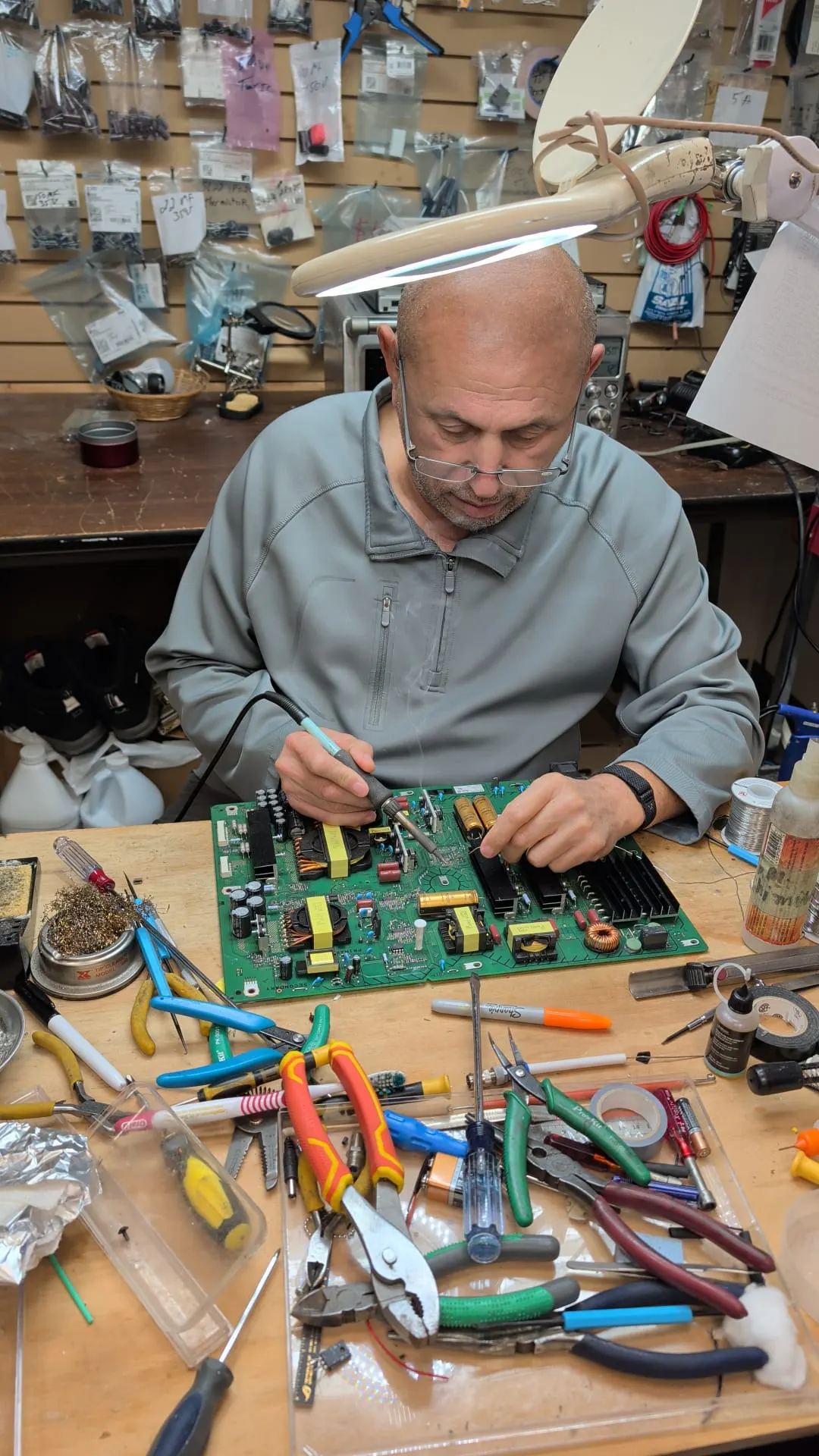 Danny, the owner of AGC Electronics Plus, at his bench in Whitby, soldering a TV power supply board under a magnifier lamp